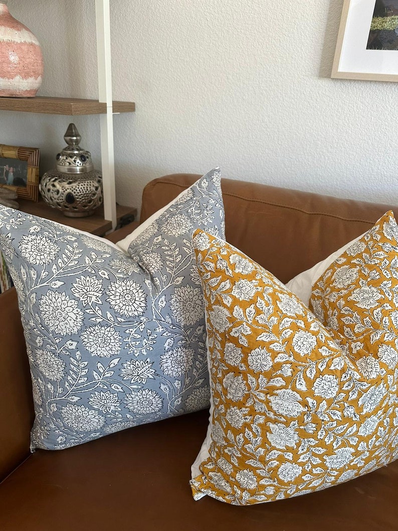 K&ouml;nnte beinhalten: Zwei quadratische Dekokissen mit einem detaillierten floralen Blockdruckmuster. Ein Kissen ist hellblau mit wei&szlig;en Blumenmustern, das andere senfgelb mit wei&szlig;en Blumenmustern. Beide Kissen haben eine R&uuml;ckseite aus wei&szlig;em Stoff.