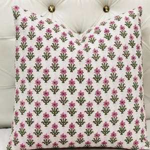 K&ouml;nnte beinhalten: Ein quadratisches Dekokissen mit wei&szlig;em Hintergrund und einem sich wiederholenden Muster aus kleinen rosa Bl&uuml;ten und gr&uuml;nen Stielen. Das florale Design ist in einem gitterartigen Layout &uuml;ber die gesamte Oberfl&auml;che des Kissenbezugs verteilt.