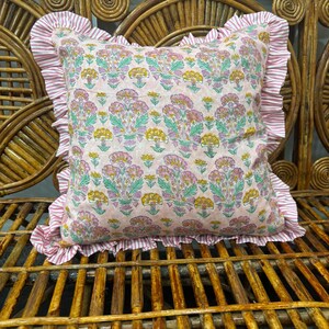 May include: A square throw cushion featuring a light pink background with a repeating floral pattern in shades of pink, green, and yellow. The cushion is bordered by a ruffled edge made of fabric with thin white and pink vertical stripes.