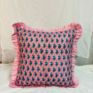 May include: A square throw pillow featuring a pink background with a repeating blue and green floral block print pattern. The pillow is finished with a ruffled border made of fabric with thin red and white vertical stripes on all four sides.