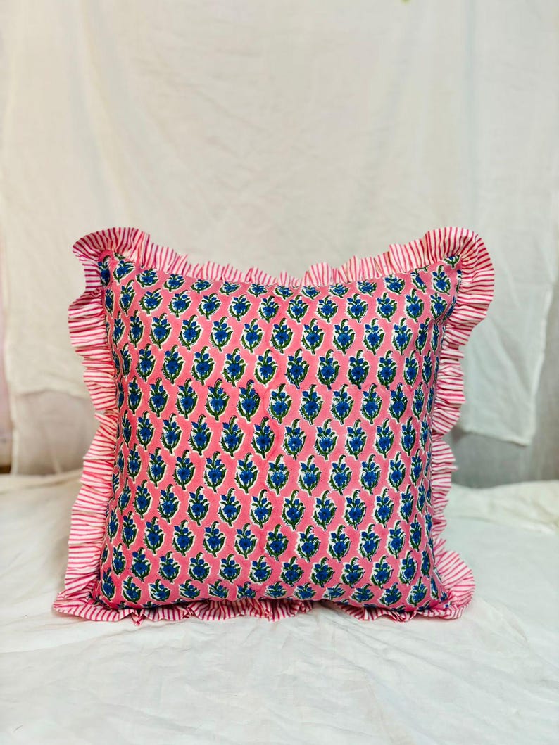 May include: A square throw pillow featuring a pink background with a repeating blue and green floral block print pattern. The pillow is finished with a ruffled border made of fabric with thin red and white vertical stripes on all four sides.