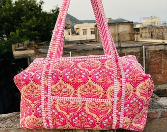 May include: A rectangular quilted duffel bag featuring a vibrant pink and cream floral block print pattern. The bag includes two long shoulder straps with a pink and white striped design, matching the trim along the edges and the central pocket.