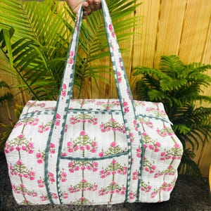 May include: White quilted tote bag with a pink floral pattern and green trim. The bag has two handles and a rectangular shape. The floral design is repeated across the bag's surface, creating a cohesive and visually appealing look.