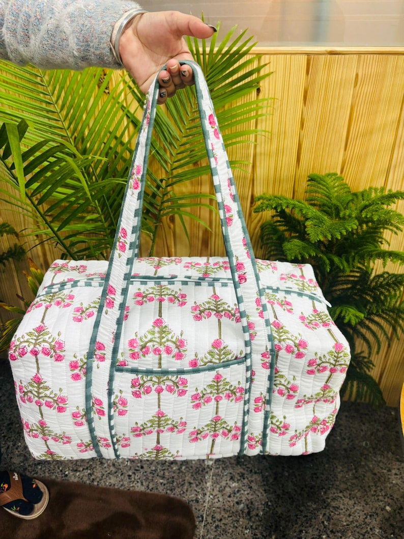 May include: White quilted tote bag with a pink floral pattern and green trim. The bag has two handles and a rectangular shape. The floral design is repeated across the bag's surface, creating a cohesive and visually appealing look.