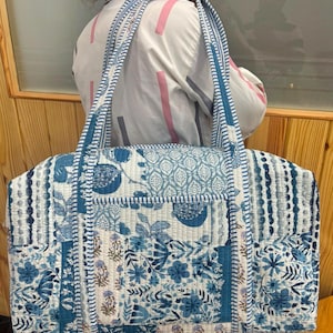 May include: A large rectangular quilted tote bag featuring a patchwork design in shades of blue and white. The fabric displays various floral, botanical, and geometric patterns. It includes two long shoulder straps and a front exterior pocket.