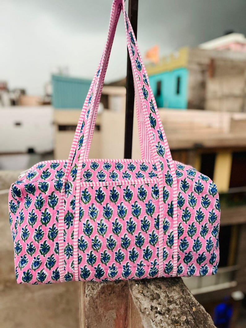 May include: Pink and blue floral patterned tote bag with matching handles. The bag features a rectangular shape with a zipper closure and a white stripe detail. The bag is made of a soft fabric.