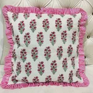 May include: A square throw pillow featuring a white base with a repeating pattern of small pink flowers and green stems. The pillow is bordered by a ruffled edge made of fabric with thin, vertical pink and white stripes on all four sides.