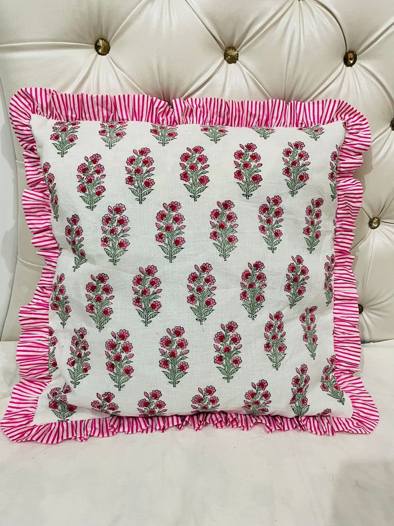 May include: A square throw pillow featuring a white base with a repeating pattern of small pink flowers and green stems. The pillow is bordered by a ruffled edge made of fabric with thin, vertical pink and white stripes on all four sides.