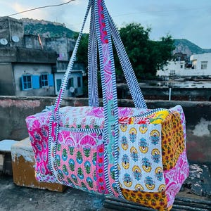 May include: A colorful, quilted duffel bag with a patchwork design. The bag features pink, yellow, and blue floral patterns, with striped and paisley accents. The handles are made of striped fabric.