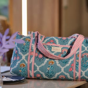 May include: A quilted tote bag with a floral pattern in teal, pink, and white. The bag features a geometric design with pink stripes and handles. The bag is designed for carrying items and has a rectangular shape.