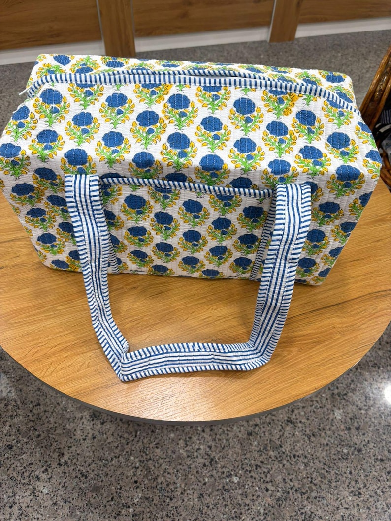 May include: A rectangular tote bag with a floral pattern of blue flowers and yellow accents on a white background. The bag has blue and white striped handles and trim. The bag is sitting on a wooden table.