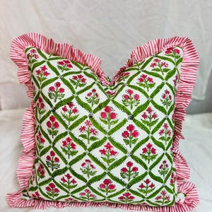 May include: A square throw pillow featuring a white base with a green diamond lattice pattern containing pink floral motifs. The pillow is bordered by a ruffled edge made of fabric with thin red and white vertical stripes throughout the trim.