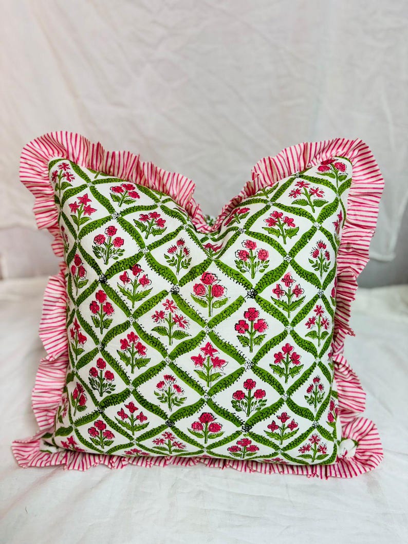 May include: A square throw pillow featuring a white base with a green diamond lattice pattern containing pink floral motifs. The pillow is bordered by a ruffled edge made of fabric with thin red and white vertical stripes throughout the trim.