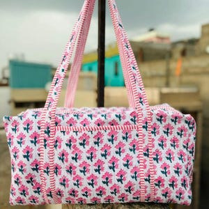 May include: A white and pink floral print tote bag with matching handles. The bag features a repeating pattern of pink flowers with green stems and leaves. The handles are striped with pink and white. The bag is designed for carrying items.