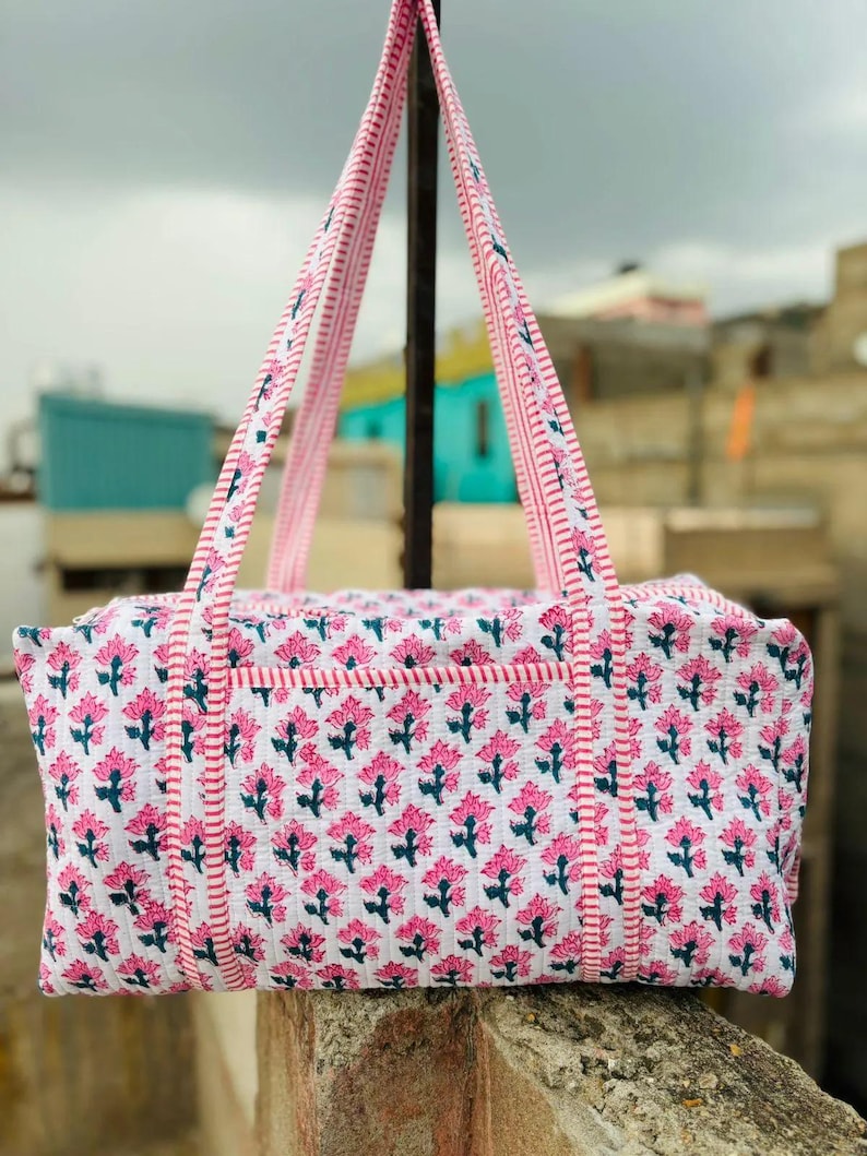May include: A white and pink floral print tote bag with matching handles. The bag features a repeating pattern of pink flowers with green stems and leaves. The handles are striped with pink and white. The bag is designed for carrying items.
