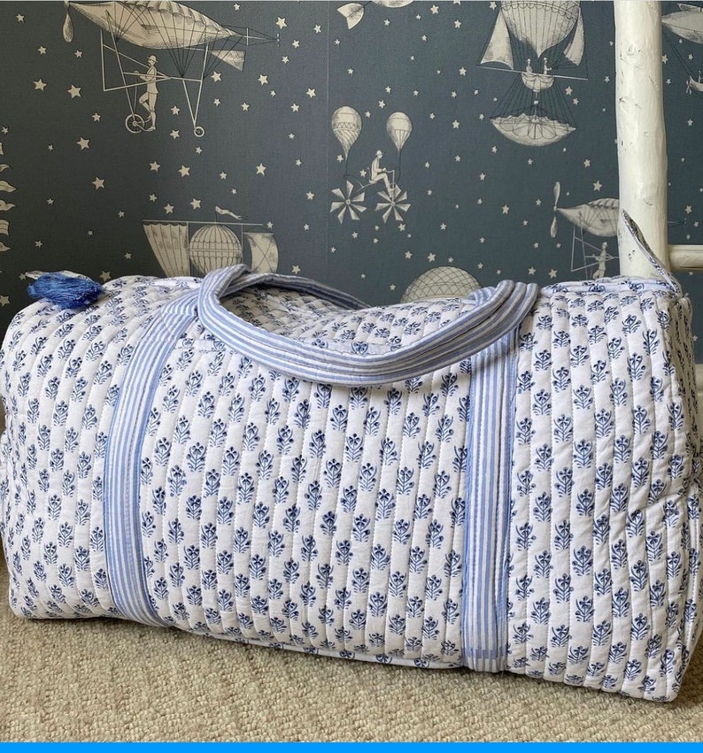 May include: A quilted duffel bag with a white background and a blue floral pattern. The bag has blue striped accents and a blue tassel. The handles are also blue and white striped. The bag is designed for travel and is made of a soft, quilted material.