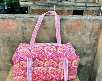 May include: A pink and beige patterned tote bag with matching striped handles. The bag features a floral design and a front pocket. The bag is sitting on a weathered surface, with a cityscape in the background.