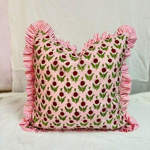 May include: A square throw pillow featuring a light pink base with a repeating pattern of dark red circular flower heads and green leaves. The pillow is bordered by a ruffled edge made of fabric with thin, alternating white and red vertical stripes.