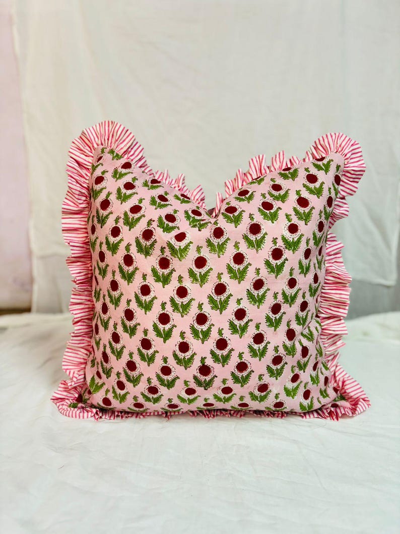 May include: A square throw pillow featuring a light pink base with a repeating pattern of dark red circular flower heads and green leaves. The pillow is bordered by a ruffled edge made of fabric with thin, alternating white and red vertical stripes.