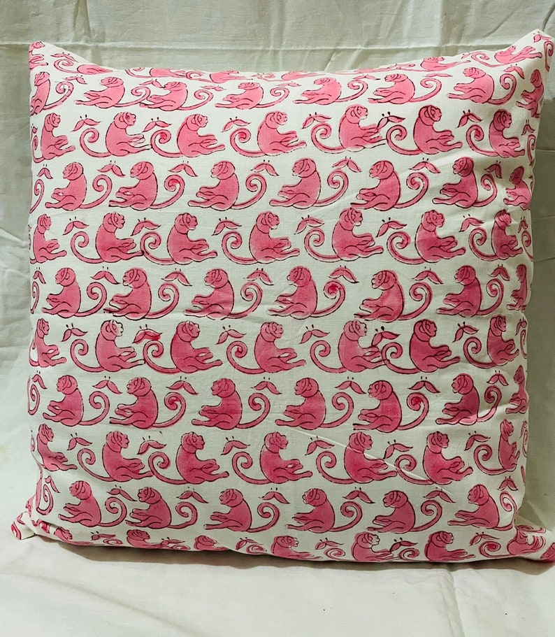 K&ouml;nnte beinhalten: Ein quadratisches Kissen mit einem cremewei&szlig;en Stoffgrund und einem sich wiederholenden Muster aus rosa Affen in sitzender Position. Jeder Affe ist im Profil dargestellt und wechselt sich mit kleinen rosa blattartigen Formen in Reihen ab.