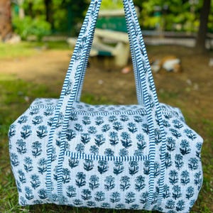 May include: A white and blue floral patterned tote bag with matching handles. The bag features a repeating floral design in shades of blue on a white background. The handles are a striped blue and white pattern.