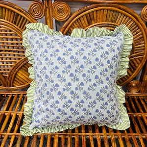 May include: A square throw cushion featuring a light blue fabric base with a repeating pattern of small blue flowers and green stems. The cushion is bordered by a ruffled edge made of fabric with thin, vertical green and white stripes.