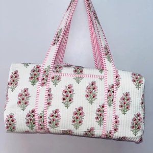 May include: White quilted tote bag with a floral pattern in pink and green. The bag features pink striped handles and trim, and a small pocket on the front. The bag is hanging against a white background.