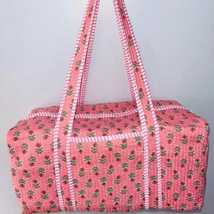 May include: Pink quilted duffel bag with floral print. The bag features matching pink handles and a white striped border. The floral pattern includes red flowers and green leaves. The bag is designed for travel or carrying items.