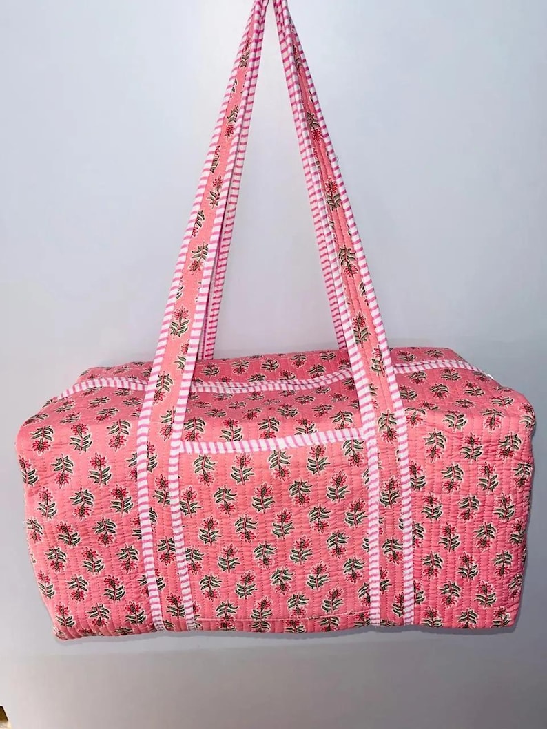 May include: Pink quilted duffel bag with floral print. The bag features matching pink handles and a white striped border. The floral pattern includes red flowers and green leaves. The bag is designed for travel or carrying items.