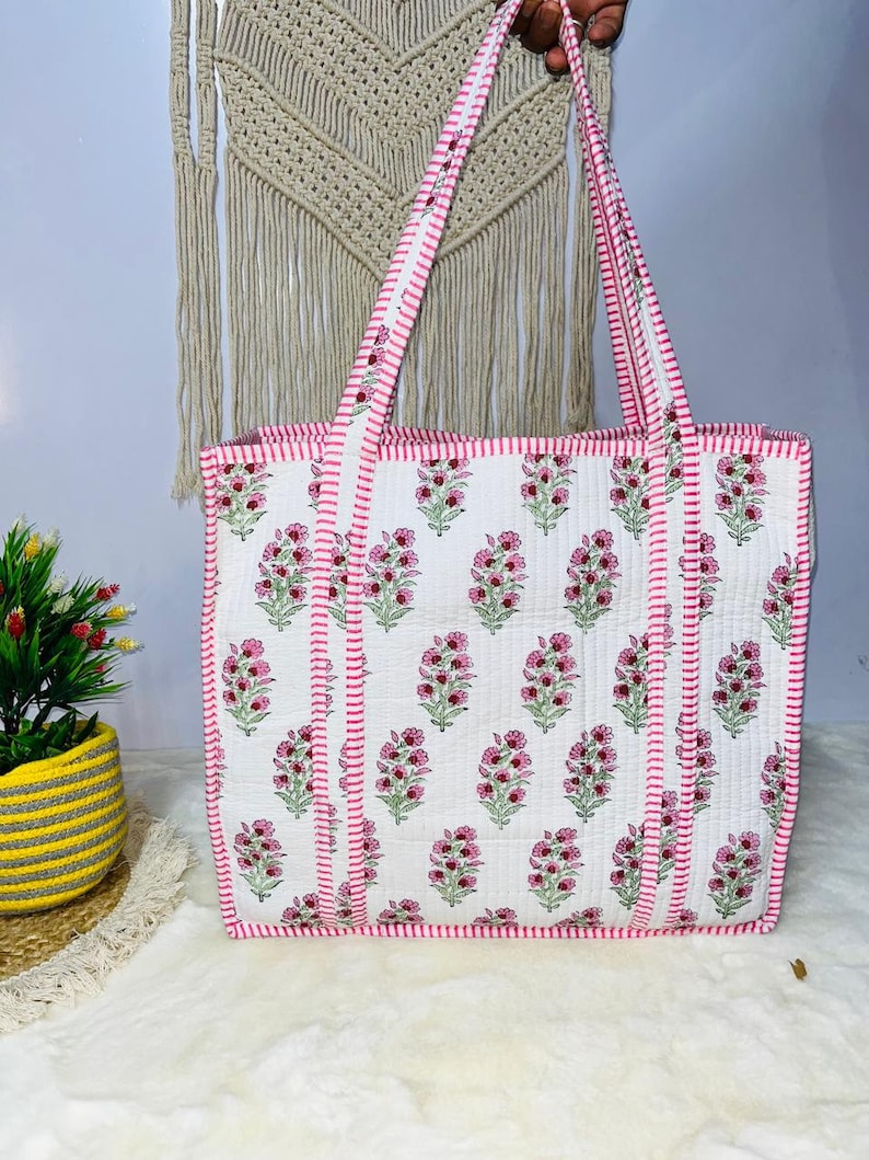 Pu&ograve; includere: Una borsa tote in cotone bianco con stampa floreale rosa e manici a righe rosa e bianche. La borsa ha un bordo cucito rosa.