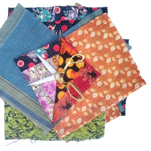 May include: A stack of colorful fabric squares with a gold pair of scissors on top. The fabric squares are made of different patterns and colors, including blue, green, orange, red, and purple.