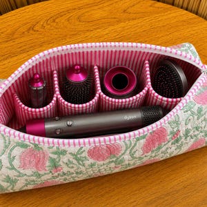 May include: A quilted fabric storage case with a pink and white striped interior and a floral exterior pattern in pink and green. The case contains a grey and pink Dyson hair styling tool and four circular hair styling attachments in individual slots.
