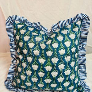 May include: A square throw pillow featuring a deep teal background with a repeating pattern of white flowers and green stems. The pillow is bordered by a ruffled edge made of fabric with a blue and white horizontal stripe pattern throughout.
