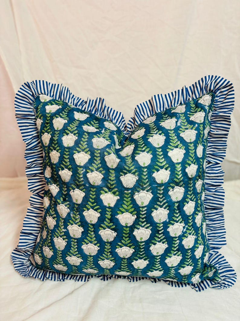 May include: A square throw pillow featuring a deep teal background with a repeating pattern of white flowers and green stems. The pillow is bordered by a ruffled edge made of fabric with a blue and white horizontal stripe pattern throughout.