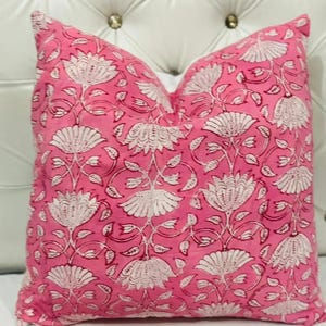 K&ouml;nnte beinhalten: Ein quadratisches Zierkissen mit einem leuchtend rosa Hintergrund und einem sich wiederholenden wei&szlig;en Blumenmuster aus stilisierten lotusartigen Bl&uuml;ten und zarten Bl&auml;ttern. Das Kissen wirkt weich und prall und ist frontal ausgerichtet.