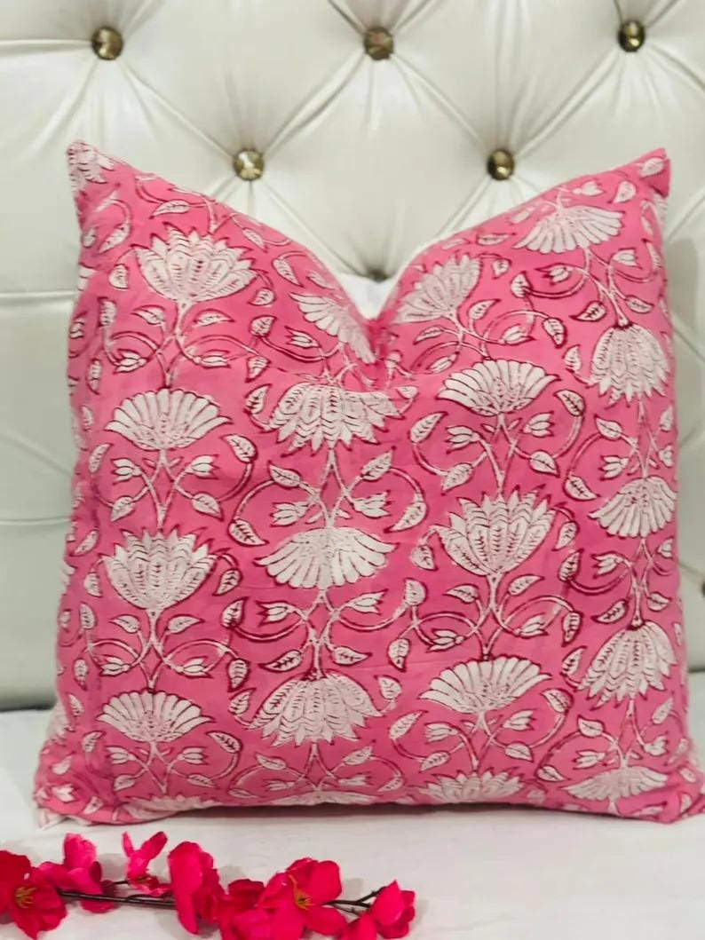 K&ouml;nnte beinhalten: Ein quadratisches Zierkissen mit einem leuchtend rosa Hintergrund und einem sich wiederholenden wei&szlig;en Blumenmuster aus stilisierten lotusartigen Bl&uuml;ten und zarten Bl&auml;ttern. Das Kissen wirkt weich und prall und ist frontal ausgerichtet.