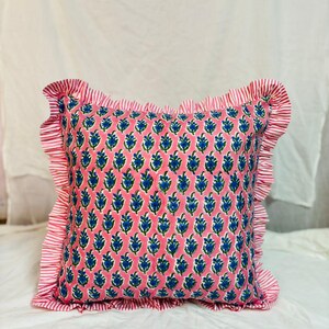 May include: A square throw cushion featuring a pink background with a repeating blue and green floral pattern. The cushion is bordered by a ruffled edge made of fabric with thin red and white vertical stripes, creating a decorative frame.