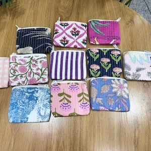Puede incluir: Una colección de pequeñas bolsas cuadradas de tela con cremalleras, cada una con patrones y colores únicos. Los diseños incluyen motivos florales, a rayas y geométricos en rosa, azul, morado y otros colores. Las bolsas están dispuestas sobre una superficie de madera.