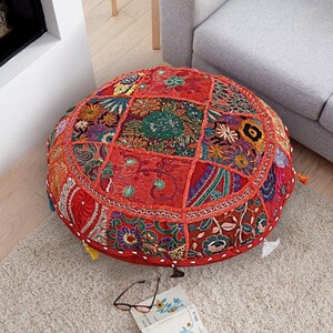 May include: A round, red, patchwork floor cushion with colorful embroidery and tassels. The cushion is made of fabric scraps sewn together in a variety of patterns and colors.