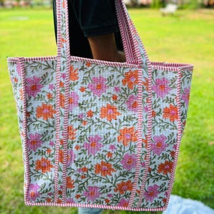 May include: A white tote bag with a floral print in shades of pink and orange. The bag has pink and white striped straps and a pink and white stitched border.