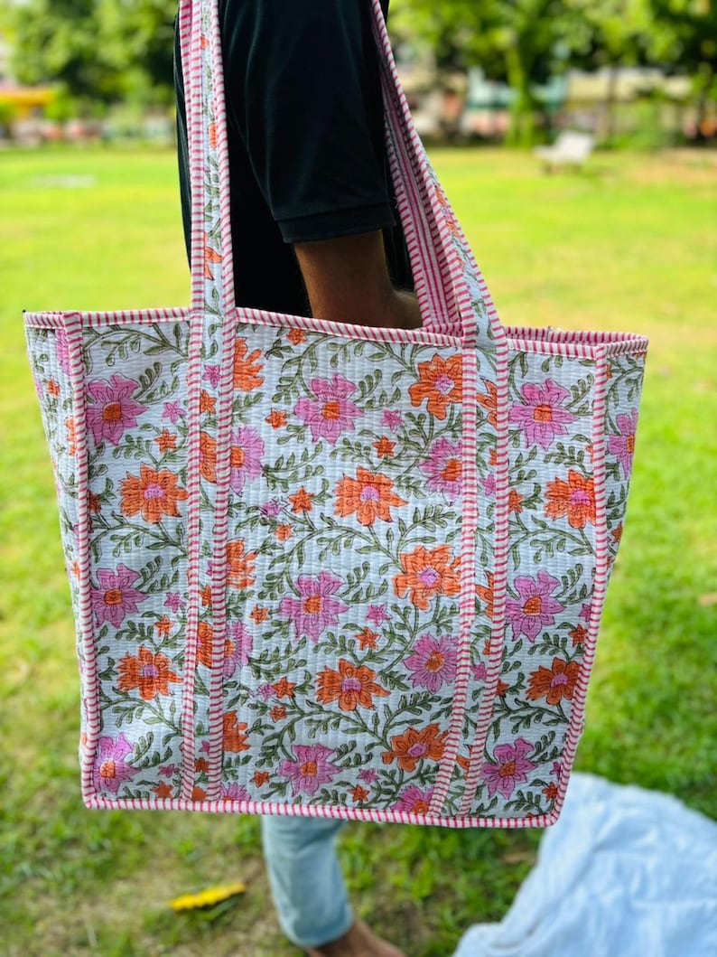 May include: A white tote bag with a floral print in shades of pink and orange. The bag has pink and white striped straps and a pink and white stitched border.