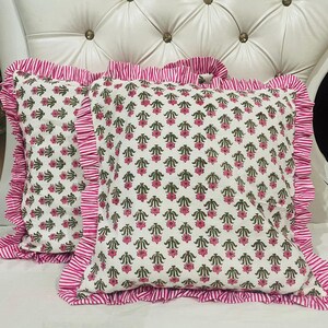 May include: Two square throw cushions featuring a white base with a repeating pattern of small pink flowers and green leaves. Each cushion is finished with a ruffled border made of fabric with thin pink and white vertical stripes along the edges.