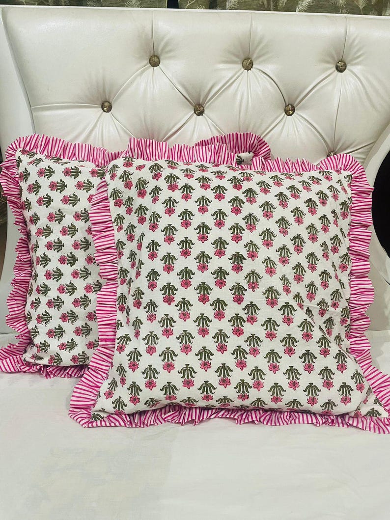 May include: Two square throw cushions featuring a white base with a repeating pattern of small pink flowers and green leaves. Each cushion is finished with a ruffled border made of fabric with thin pink and white vertical stripes along the edges.