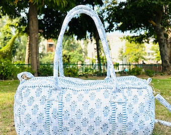 May include: A white and blue patterned duffel bag with a top handle and a shoulder strap. The bag is made of a soft, quilted fabric and has a floral design.