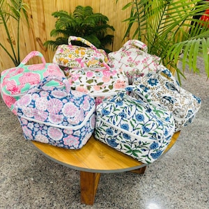 May include: A collection of quilted cosmetic bags with floral patterns in various colors, including pink, blue, and green. Each bag features a top handle and a zippered closure. The bags are arranged on a round wooden table.