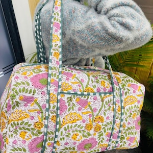 May include: A floral print duffel bag with a white background, featuring pink and yellow flowers, green leaves, and a green and white checkered strap. The bag has a rectangular shape with a pocket on the side.