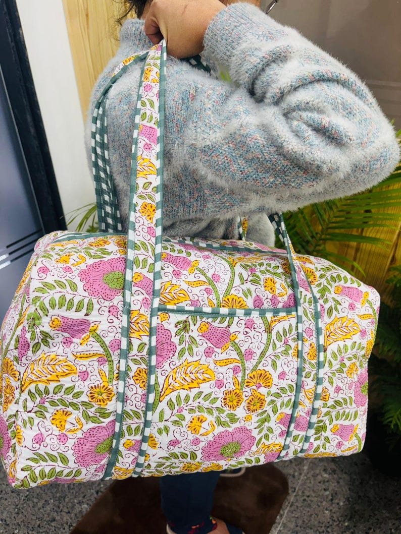 May include: A floral print duffel bag with a white background, featuring pink and yellow flowers, green leaves, and a green and white checkered strap. The bag has a rectangular shape with a pocket on the side.