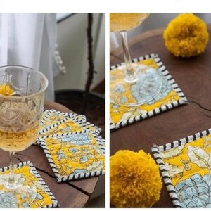 May include: Set of six yellow and blue floral coasters with black and white trim. The coasters are made of fabric and have a quilted design. Two glasses of wine are sitting on the coasters.