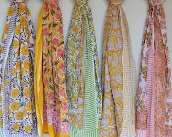 May include: Five colourful cotton scarves with floral patterns in shades of yellow, pink, green, and white. The scarves are hanging from a metal rod.