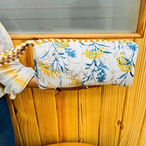 May include: A rectangular quilted pouch featuring a white base with a botanical print of yellow mimosa flowers and blue leaves. The pouch includes a matching striped wrist strap and a zipper closure along the top edge with a quilted texture.